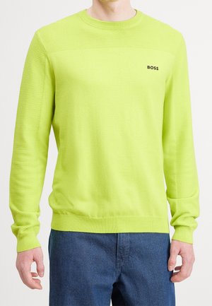 Homme portant un pull en maille vert citron à manches longues avec des manches côtelées et un logo "BOSS" noir sur la poitrine, associé à un pantalon bleu foncé.