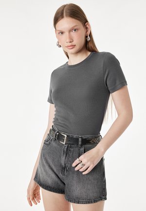 Giovane donna con capelli lunghi che indossa orecchini a cerchio in argento, maglietta aderente grigio scuro, shorts di jeans neri a vita alta e cintura nera con borchie.