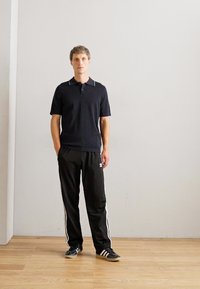 Un homme se tient devant un mur blanc uni, portant un polo noir, un pantalon de jogging noir Adidas avec des rayures blanches et des baskets noires Adidas.