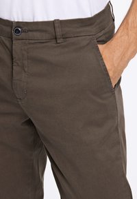 Sima textúrájú khaki chino nadrág, oldalsó zsebekkel és gombos záródással. A varrás részletei láthatók az illesztések mentén.