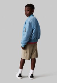 Giubbotto bomber azzurro con polsini a costine, pantaloni cargo beige, calzini bianchi e sneakers con dettagli neri. Vista posteriore, modello in posizione laterale.