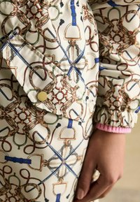 Balta auduma ar rakstiem, kas attēlo jāšanas tēmas grafikā zilā, zaļā un brūnā krāsā. Ietver spiedpogu un elastīgu aproci rozā krāsā.