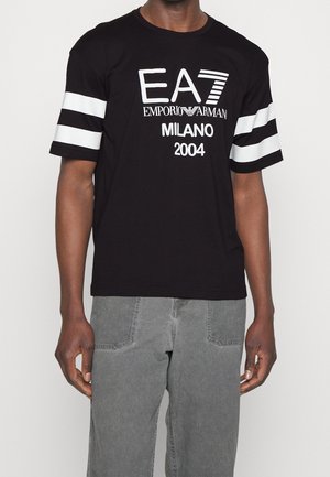 Sort kortærmet skjorte med hvide striber på ærmerne og hvid tekst "EA7 Emporio Armani Milano 2004", båret med grå bukser.