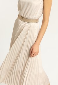 Spódnica plisowana z jasnobeżowej tkaniny o teksturowanym połysku. Uzupełniona szerokim złotym paskiem jako detal.