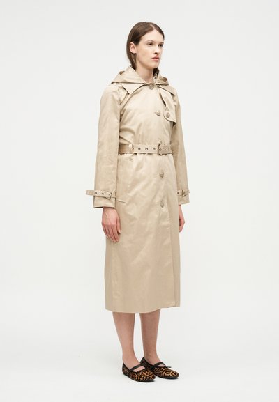 Femme debout portant un trench-coat beige mi-long ceinturé et des chaussures plates à imprimé léopard sur un fond blanc uni.
