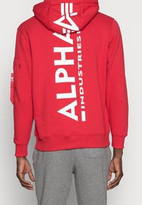 Červená mikina s kapucí vyrobená z bavlny s bílým logem "ALPHA INDUSTRIES" na zadní straně. Obsahuje kapuci na šňůrku a žebrované manžety. Pod ní jsou vidět šedé kalhoty.