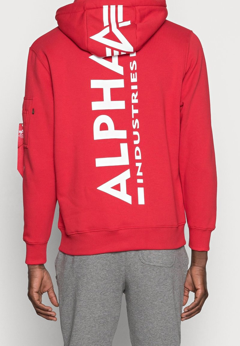 Červená mikina s kapucí vyrobená z bavlny s bílým logem "ALPHA INDUSTRIES" na zadní straně. Obsahuje kapuci na šňůrku a žebrované manžety. Pod ní jsou vidět šedé kalhoty.