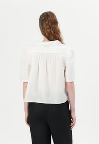 Hvit kortermet bluse med krage, med et samlet bakdetalj og subtil tekstur. Kombinert med svarte bukser, vist fra baksiden.