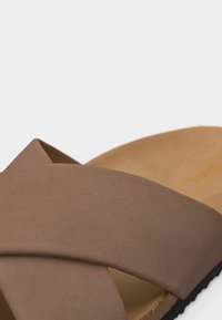 Καφέ σανδάλι με φαρδιές, διασταυρούμενες λουρίδες και επενδυμένο πέλμα, εμφανίζεται σε κοντινή λήψη πάνω σε ανοιχτό φόντο.