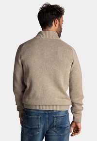 Beige gebreide pullover met een hoge geribbelde kraag en geribbelde manchetten. Draagt met blauwe denim jeans, wat zorgt voor een ontspannen pasvorm en textuur.