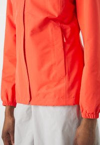 Veste imperméable orange vif avec des poignets élastiques, dotée d'une poche latérale et d'un tissu lisse et léger. Gros plan sur le bras et le torse.