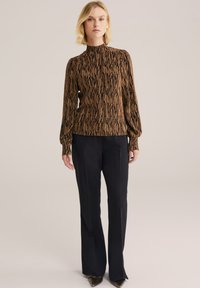 Tshirt lungo a maniche con pattern marrone e collo alto, abbinato a pantaloni neri. Il top presenta un design texture e fluido con accenti sottili.