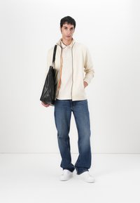 Giacca beige con zip e accenti arancioni, indossata sopra una camicia chiara, abbinata a jeans blu e sneakers bianche, mentre tiene una borsa a tracolla nera.