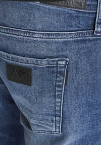 Vaqueros de mezclilla azul que presentan un bolsillo trasero con una etiqueta de cuero negro en relieve con las letras "AM". Detalles de costura visibles en toda la tela.