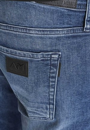 Plave traperice s stražnjim džepom na kojem se nalazi crna kožna oznaka utisnuta s "AM." Vidljive detalje šivanja kroz cijeli materijal.