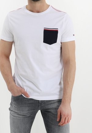 Mężczyzna ubrany w biały T-shirt z czarną kieszenią na piersi obramowaną czerwono-białymi paskami, w połączeniu z szarymi dżinsami, stoi z jedną ręką w kieszeni.