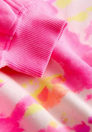 Közeli kép egy rózsaszín bordázott mandzsettáról, amely egy rózsaszín, sárga és narancssárga foltokkal tarkított batikolásos anyagon van.