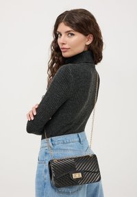 Borsa a tracolla nera trapuntata con catena dorata, caratterizzata da un motivo a zigzag e chiusura rettangolare. Indossata sopra una gonna in denim blu.