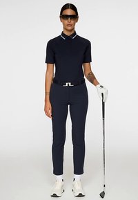 Marinblå golfoutfit med kortärmad pikétröja med vita detaljer, figurnära byxor, vita handskar och solglasögon; håller i en golfklubba.