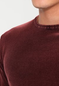 Chemise en coton bordeaux à manches longues avec col rond et col côtelé, présentant un tissu lisse légèrement texturé.