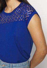Top azul de manga corta con un yugo de malla estampada. Textura suave con diseño de zigzag y escote redondeado, combinado con pantalones cortos de mezclilla.