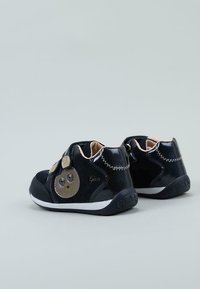 Zapatillas para niños negras con parte superior suave y texturizada, presentan un diseño de personaje sonriente, correa de Velcro y suela de goma blanca.