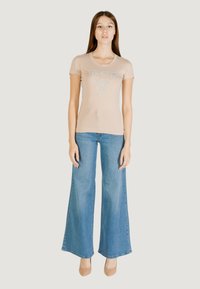 T-shirt aderente beige con logo "GUESS" in argento, abbinata a jeans in denim a gamba larga blu chiaro. Modella indossa tacchi beige. Sfondo semplice.