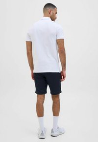 Camiseta polo blanca, de manga corta, con dobladillo recto. Llevada con pantalones cortos azul marino. El modelo usa calcetines blancos y zapatillas deportivas grises. Diseño minimalista.