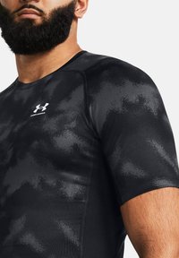 Zwart sportshirt met korte mouwen, voorzien van een camo-patroon en het Under Armour-logo op de borst. Gemaakt van rekbaar, glad materiaal.