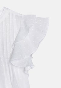 Blusa bianca con tessuto testurizzato, caratterizzata da morbide maniche a balze, sottili punti ricamati e righe verticali che scendono lungo il corpo.