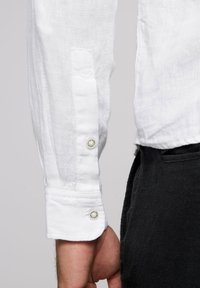 Camisa de lino blanca con mangas largas, que presenta puños abotonados y una tela texturizada. La camisa se combina con pantalones oscuros.