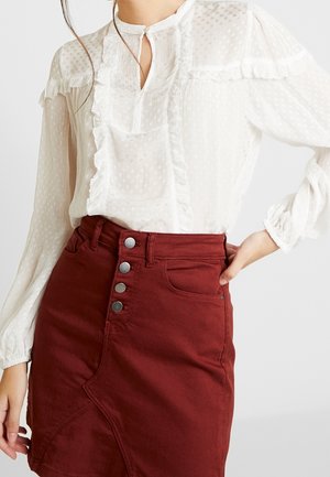 Blouse blanche texturée à manches longues avec volants, associée à une jupe rouge taille haute boutonnée, main posée sur la hanche.