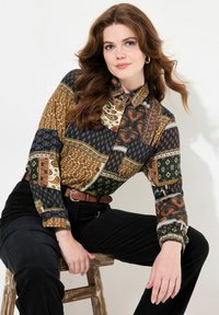 Camisa de manga larga con botones que presenta un diseño de patchwork en dorado, negro y verde con varios patrones. Combinada con pantalones negros y un cinturón marrón.