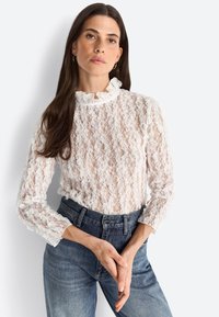Top in pizzo bianco con scollatura alta a forma di scallop e maniche lunghe, caratterizzato da un motivo intricato e una leggera trasparenza. Jeans blu a vita alta.