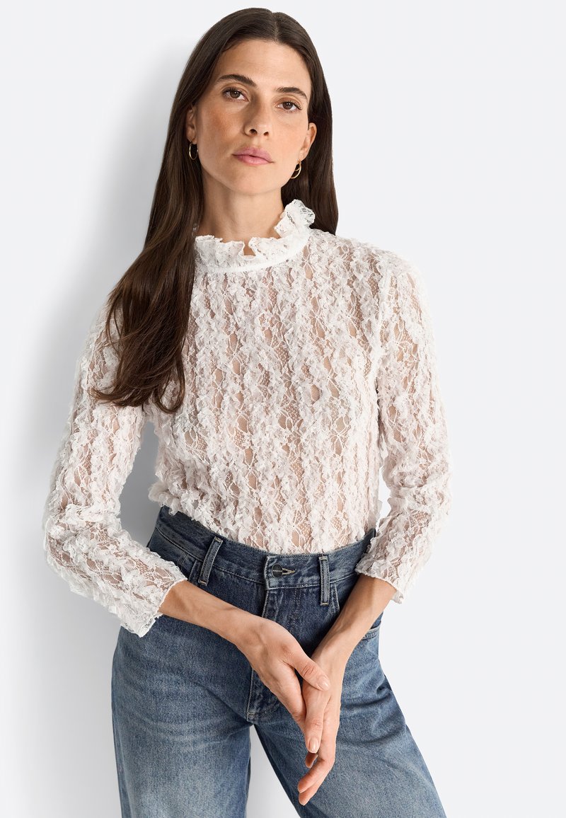 Top in pizzo bianco con scollatura alta a forma di scallop e maniche lunghe, caratterizzato da un motivo intricato e una leggera trasparenza. Jeans blu a vita alta.
