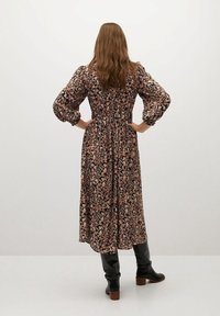 Robe noire à fleurs avec manches bouffantes, taille cintrée et jupe longueur cheville. Le tissu présente un motif floral multicolore. Portée avec des bottes noires.