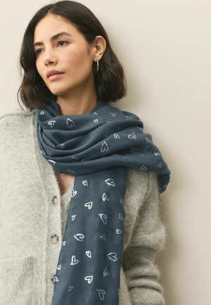 Woman with short dark hair wearing silver hoop earrings, a soft gray buttoned cardigan, and a blue scarf with silver heart patterns.