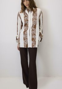 Smeđi i bijeli košulja s uzorkom s ovratnikom, ukrašena čipkastim cvjetnim detaljima, u kombinaciji s tamnosmešim hlačama širokih nogavica.
