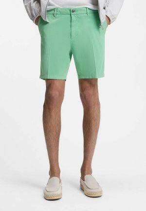 Uomo che indossa pantaloncini su misura verde chiaro, scarpe bianche senza lacci e una giacca leggera con le mani in tasca, in piedi contro uno sfondo bianco.