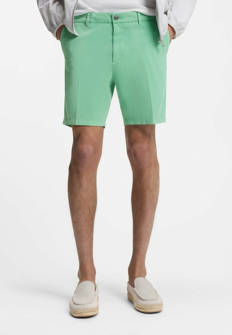 Mann trägt hellgrüne, maßgeschneiderte Shorts, weiße Slip-On-Schuhe und eine leichte Jacke, die Hände in den Taschen, steht vor weißem Hintergrund.