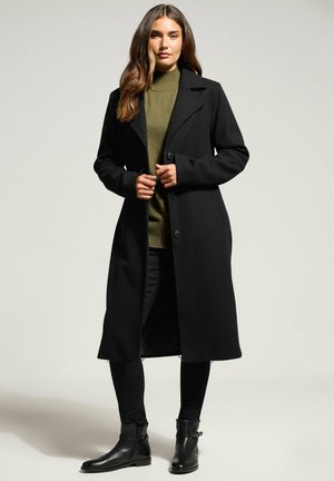 Donna con lunghi capelli castani che indossa un lungo cappotto nero, un maglione a collo alto verde oliva, pantaloni neri e stivali neri alla caviglia, in piedi su uno sfondo neutro.