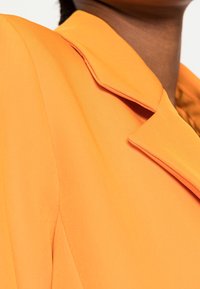Blazer naranja con cuello estructurado y tela lisa, con un detalle de un solo botón cerca de la solapa y costuras limpias.