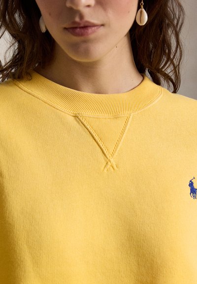 Κοντινό πλάνο ενός ατόμου που φοράει κίτρινο πουλόβερ με στρογγυλή λαιμόκοψη και μπλε κεντημένο λογότυπο, με ένα μόνο σκουλαρίκι κοχύλι στο αριστερό αυτί.