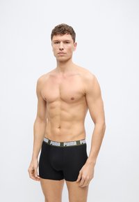 Νέος μυώδης άνδρας με κοντά σγουρά μαλλιά που φοράει μαύρα ανδρικά εσώρουχα Puma στέκεται μπροστά σε ένα απλό φωτεινό φόντο.