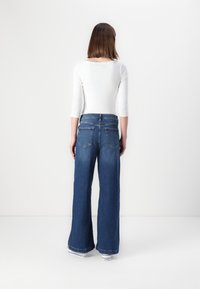 Lange, weit geschnittene Jeans mit dunkler Wäsche, fünf Taschen und hoher Taille. Kombiniert mit einem figurbetonten weißen Langarmoberteil und weißen Sneakers.