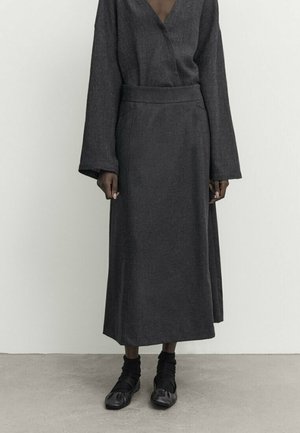 Jupe longue gris foncé et haut assorti à manches longues avec manches larges, porté avec des chaussettes noires et des chaussures noires souples sur un sol en béton.