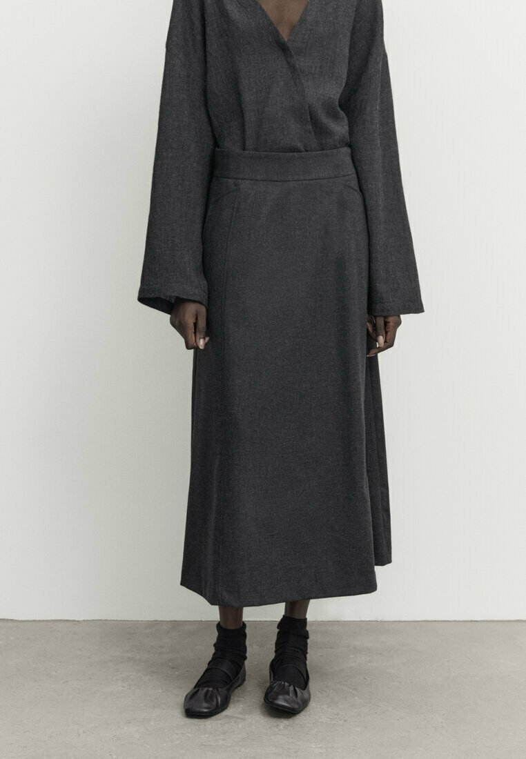 Jupe longue gris foncé et haut assorti à manches longues avec manches larges, porté avec des chaussettes noires et des chaussures noires souples sur un sol en béton.