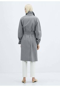 Trench coat gri cu centură, guler înalt, mâneci strânse și buzunare laterale, asortat cu pantaloni albi scurtați și pantofi bej.