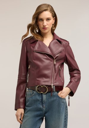 Frau trägt eine bordeauxrote Leder-Bikerjacke, blaue Jeans, goldene Creolen und einen braunen Gürtel mit einer goldenen Doppelring-Schnalle, steht mit einer Hand in der Tasche.