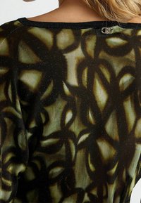 Musta ja vihreäkuvioinen pusero, jossa on teksturoitu, läpikuultava kangas. Takana lukko-ominaisuus ja resorilla varustettu kaula-aukko.